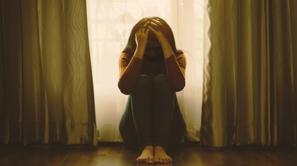 Sad woman sitting on floor in front of windows with her head in her hand