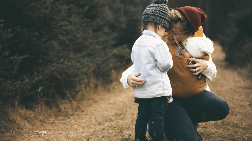 mother kneeling outside carrying infant in a wrap with arm around her other child